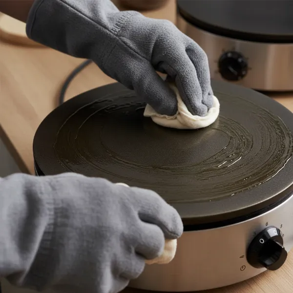 Hand applying a thin layer of vegetable oil on a hot crepe maker plate with a greasing pad.