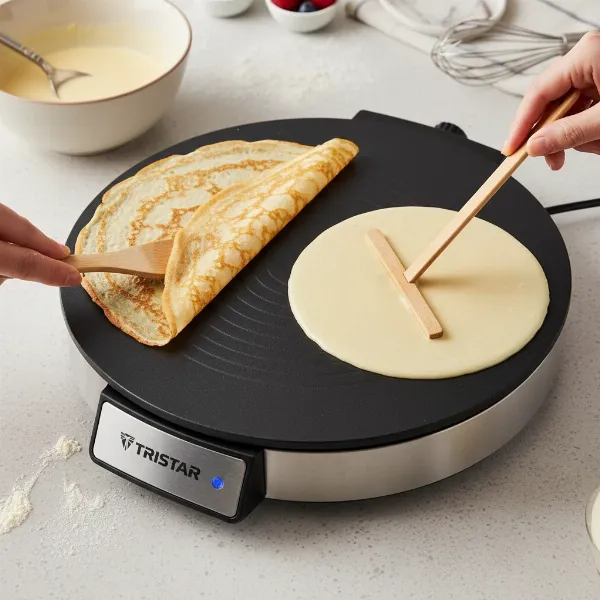 Tristar crepe maker with a wooden T-stick spreading batter and a wooden spatula flipping a crepe.